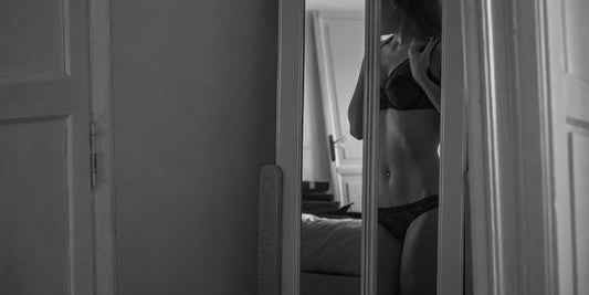  "Black and white image of a woman in lingerie looking at her reflection in the mirror, capturing quiet self-confidence."